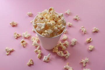Popcorn in a paper cup. The cup is overflowing and the popcorn is scattered everywhere. Pink background.