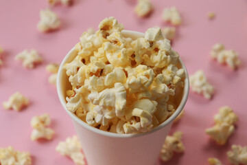 Popcorn in a paper cup. The cup is overflowing and the popcorn is scattered everywhere. Pink background.