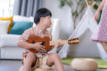 Young child with Down syndrome concentrating while playing a ukulele, sitting indoors in a warm...