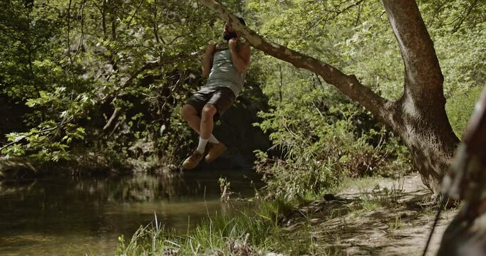 Man performs pull-ups on a tree branch by the river while camping. He enjoys the outdoors and exercises amidst nature.
