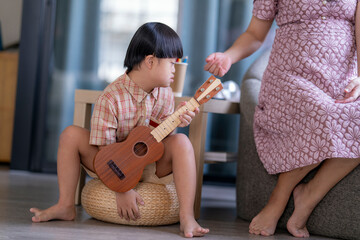 Young child with Down syndrome concentrating while playing a ukulele, sitting indoors in a warm...