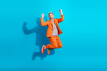 Obraz premium Charismatic gentleman in stylish orange outfit jumping with excitement against a vibrant blue background symbolizing joy