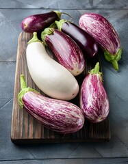  Various fresh white, striped and purple eggplants on wooden cutting board on gray wooden table. Generated image