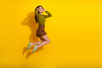 Young woman jumping joyfully on a vibrant yellow backdrop, wearing trendy khaki and green casual outfit with glasses