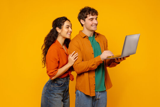 Positive couple using laptop, man and woman looking at computer screen and smiling, surfing on Internet, standing over orange background - Powered by Adobe