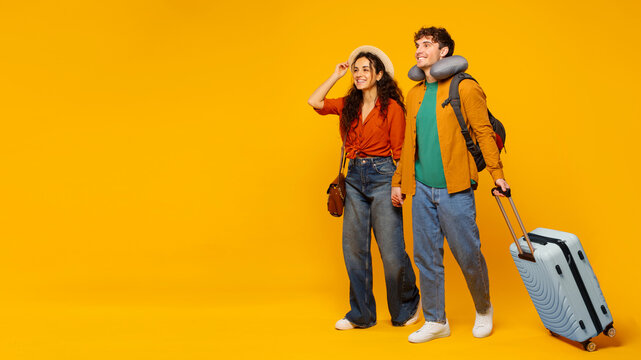 Travel offer. Happy European couple walking with suitcase and looking at copy space on orange background, full length. Spouses going to vacation together