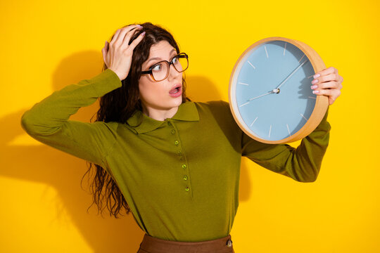 Surprised young woman holding a blue clock against yellow background depicting urgency and punctuality