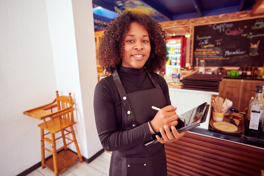Young cheerful waitress wearing black apron using digital tablet and stylus pen taking orders in cafe, looking at camera - Powered by Adobe