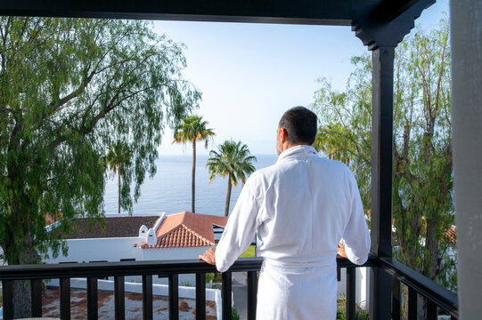A man in a white robe stands on a balcony overlooking the ocean. The scene is peaceful and serene, with the man enjoying the view and the calming sound of the waves