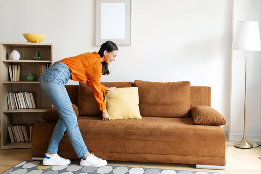 Woman carefully arranging and fluffing throw pillows on sofa, taking pride in her neat and stylishly decorated living room, full length