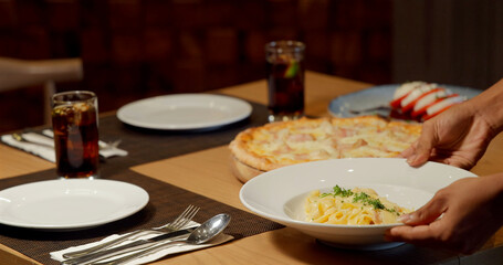 Placing pasta on table set with pizza and drinks. Cozy dining scene in restaurant. Italian cuisine, meal preparation, relaxing atmosphere.