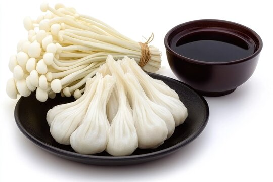 Steamed dumplings arranged on a dark plate, accompanied by a bundle of enoki mushrooms and a small bowl of dark sauce, all set against a bright white background