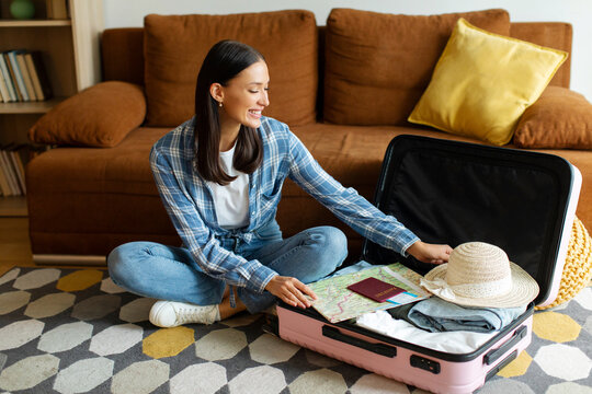 Happy European woman sitting on floor and packing clothes into open suitcase, getting ready for trip, preparing luggage for journey