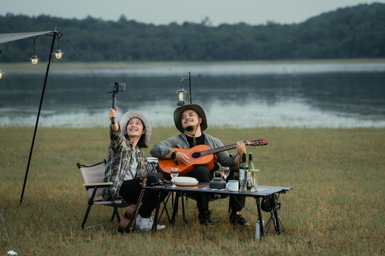 A man and a woman are sitting on a lawn with a guitar. The man is holding a camera. camping outdoor lifestyle concept. - Powered by Adobe