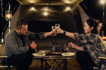A man and a woman are sitting at a table with wine glasses and eating food. They are both smiling and raising their glasses in a toast. camping outdoor lifestyle concept.