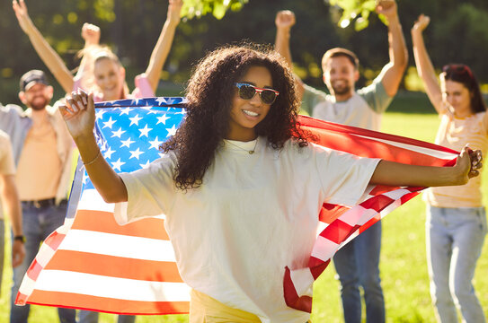Portrait of happy african american woman holding usa flag in hands celebrating Independence Day standing in summer park outdoors with smiling people friends in background. Patriotism concept.