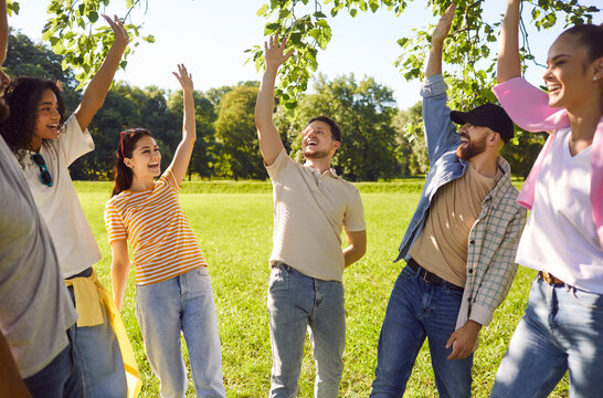 Portrait of a group of excited happy young friends students having fun hanging together outdoors in the summer park. People standing in a circle raising their hands up enjoying meeting.