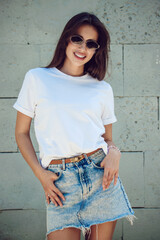 Young Woman In A White T-Shirt And Denim Skirt Posing Against Concrete Wall