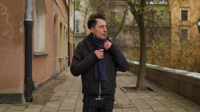 A man underdressed for the weather gets caught in the cold on a winter day. He adjusts his thin jacket and scarf, trying to stay warm as he was not prepared for the chilly wind.
