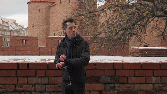 A man is waiting for someone on a historic city street in winter. He paces back and forth, checking the time on his watch and looking at his phone, possibly early for a date or a meeting.