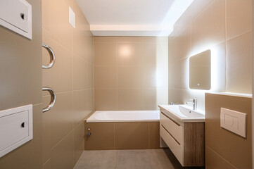 Modern bathroom with beige tiles, a white bathtub, vanity with illuminated mirror, and heated towel...