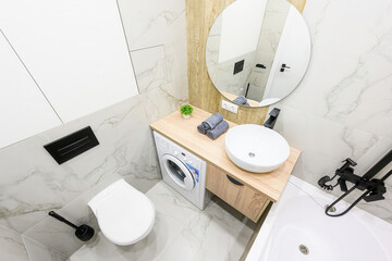 Modern bathroom with marble tiles, round mirror, wooden vanity, white sink, washing machine, and...