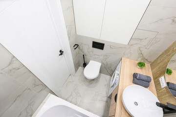Bright, modern bathroom with white marble-patterned tiles, a white toilet, sink, and wooden...