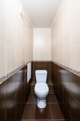 narrow tiled bathroom featuring a white toilet. The walls are half brown and beige tiles with a...