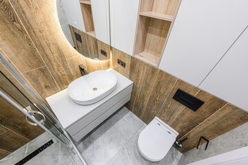 Modern bathroom with wood-look tile, white sink, toilet, and cabinet. Features a round lighted...