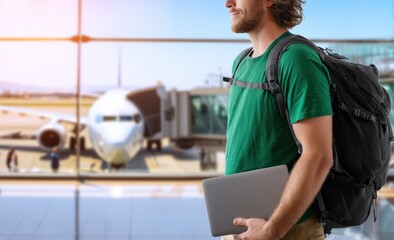 Freelancer waiting for boarding holding a laptop in his hand at the airport, with a large airplane and a jet bridge in the background