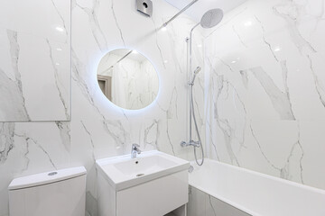 bright, modern bathroom featuring white marble-patterned tiles, a round LED mirror, and white...