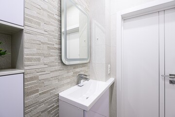 bright, modern bathroom features textured tile walls, a white vanity with a sink, and a glowing-edge mirror. A white door is visible on the right side of the frame