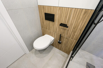 Modern bathroom featuring a toilet, tiled walls (grey, wood-like), and a shower with a black frame....