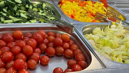 Fresh vegetables at a salad bar: cherry tomatoes, cucumbers, peppers, and lettuce in metal trays