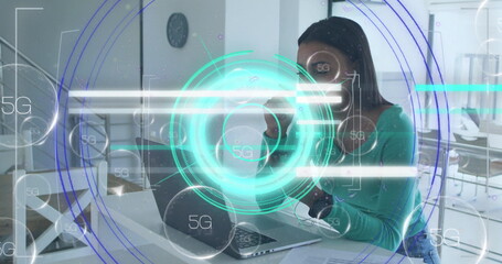 Holding mug, mid adult woman typing on silver laptop at open-plan office, with 5G interface