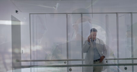 Using smartphone senior doctor in white coat speaking through glass partition at hospital corridor