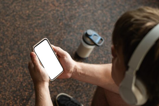 Young sporty man holding mobile phone with empty screen, while taking break in between exercises in gym. Mock up. Close up.