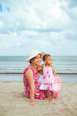 Asian mom and toddler girl in beach dresses having fun together during vacation at a tropical beach on a summer day with blue ocean and cloudy sky on the background