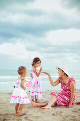 Asian mother and her two little children in beach dresses having fun together during vacation at a tropical beach on a summer day with blue ocean and cloudy sky on the background