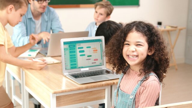 American student looking at camera and learning prompt in STEM technology class while group of diverse student and teacher doing mind mapping. Smart child learning coding software program. Pedagogy.