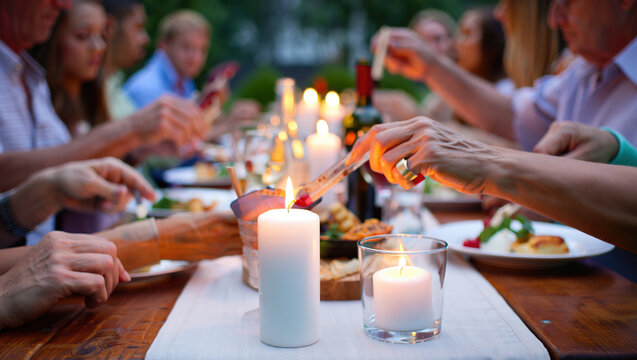 close-up of an outdoor dinner, a table covered with a white tablecloth, with various snacks, glasses, bottles and lit candles. The hands of people sitting at the table are visible. - Powered by Adobe