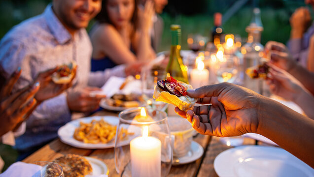 close-up of an outdoor dinner, a table covered with a white tablecloth, with various snacks, glasses, bottles and lit candles. The hands of people sitting at the table are visible. - Powered by Adobe