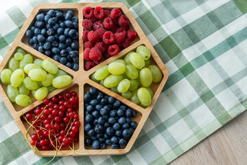 Mixed fresh berries and grapes arranged in hexagonal wooden serving tray on green checkered kitchen...