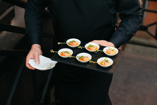 Waiter serving gourmet shrimp appetizers at event. A waiter in black uniform carries a tray of shrimp appetizers on skewers in small white bowls, ready for guests.