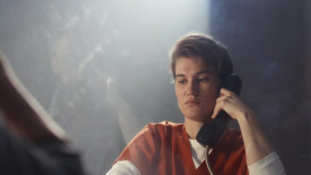 Young short-haired female inmate in orange uniform holding phone and speaking with detective through glass during interrogation in non-contact prison visitation room
