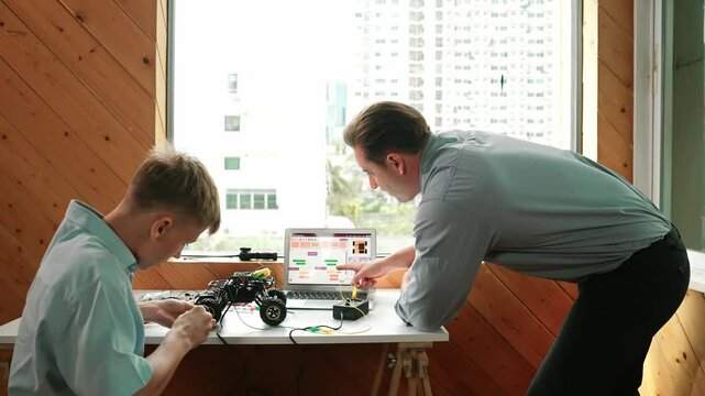 Teacher help student fixing car model and giving advise for programming system or coding prompt while high school boy listen and use equipment to repair robot at STEM classroom. Back view. Edification