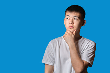 Portrait of handsome asian guy deep in thought, standing with chin in hand against blue background