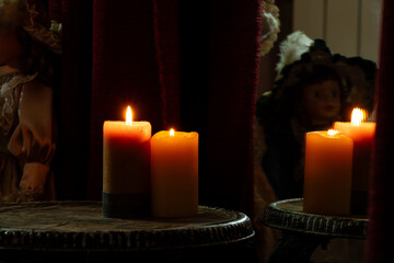 Two burning candles are standing on chairs and behind a doll and near a mirror and in the reflection of the mirror candles are visible in a room in the dark