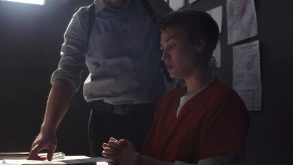 Assertive investigator pointing to photo evidence and case documents, interrogating handcuffed female inmate in prison uniform sitting at table and denying her guilt in detention room