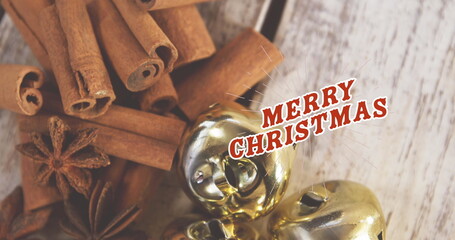 Obraz premium Displaying cinnamon sticks, anise pods, golden bells on wood surface with red Merry Christmas text